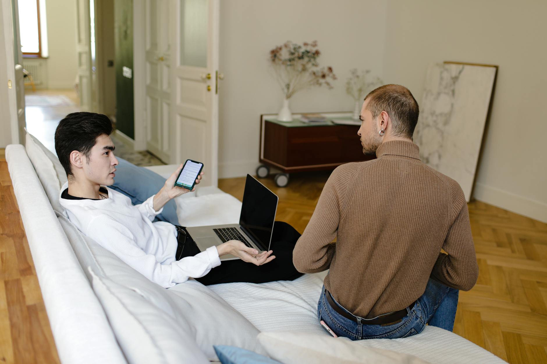 a man in white sweater holding his phone while talking to the man in brown knitted sweater